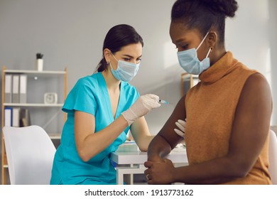 Female Doctor medical worker in blue uniform and face mask making vaccination injection against coronavirus infection for black woman patient. Vaccinating against COVID-19 infection during pandemic