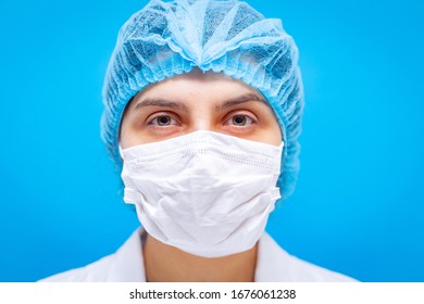 Female Doctor In Medicine Mask, Hair Cap And White Gown Portrait On A Blue Background Closeup Shot. Shallow Depth Of Field. Coronavirus, Pandemia And Medical Care Concept.