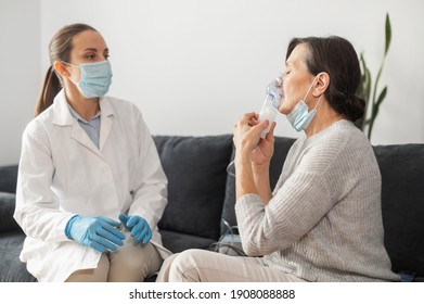 A Female Doctor, Nurse Wearing A Medical Gown And A Face Mask, Puts An Oxygen Mask On Sick Senior Patient At Home During Pandemic. Healthcare And Medicine Concept