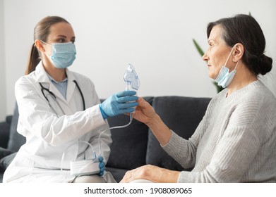 A Female Doctor, Nurse Wearing A Medical Gown And A Face Mask, Puts An Oxygen Mask On Sick Senior Patient At Home During Pandemic. Healthcare And Medicine Concept