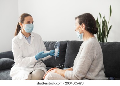 A Female Doctor, Nurse Wearing A Medical Gown And A Face Mask, Puts An Oxygen Mask On Sick Senior Patient At Home During Pandemic. Healthcare And Medicine Concept