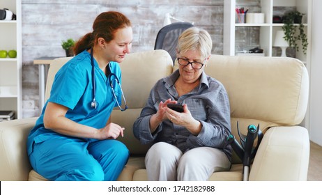 Female doctor in nursing home sitting on the couch teaching senior woman to use smartphone
