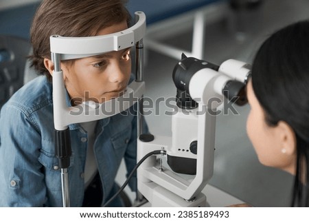 Female doctor ophthalmologist doing basic eye examination of cute boy in modern clinic. Caucasian female optician checking on kid patient vision eyesight. Myopia glaucoma prevention concept