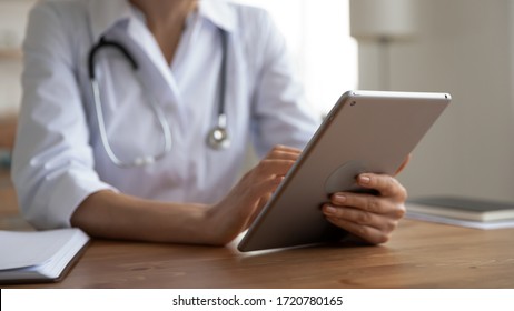 Female doctor physician wearing white coat holding modern digital tablet computer in hands using healthcare medical tech app working in software web program at workplace in hospital. Close up view.