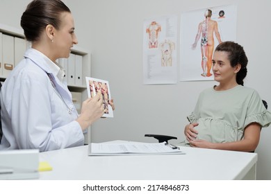 Female Doctor Pointing At Ultrasound Image And Presenting Information To Pregnant Woman During Her Visit At Hospital