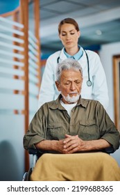 Female Doctor Pushing Worried Senior Patient In Wheelchair Though Hallway In The Hospital. Focus Is On Man.