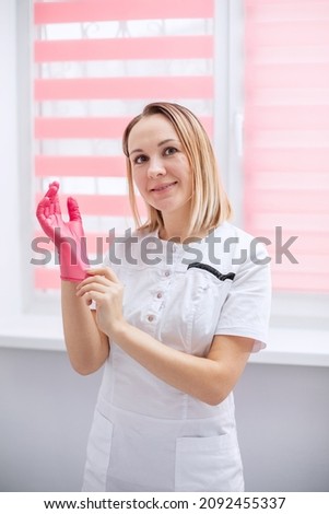 A female doctor puts on a latex glove before work. A beauty master in a white coat in the office looks at the camera and smiles. Portrait of a young woman in light colors. Vertical photo.