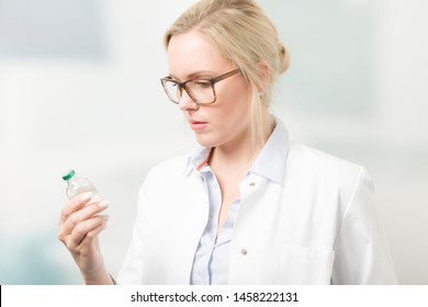 Female Doctor Is Reading Label Of Serum Bottle With Clinic Room In Background