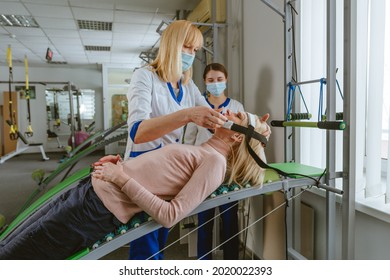 Female Doctor Rehabilitologist Working With Female Patient On The Special Rehabilitation Device At Rehabilitation Center Gym. Doctor And Nurse Wearing Protective Face Masks.