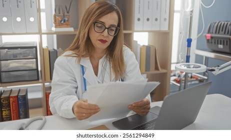 Female doctor reviewing paperwork in a clinic office while seated at a desk with laptop and medical equipment - Powered by Shutterstock