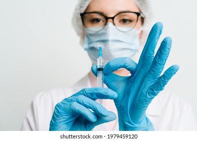 Female Doctor Or Scientist In White Medical Gown, Blue Gloves, Green Cap And Mask Holds A Syringe In Hands On White Background. She Is Ready To Give An Injection