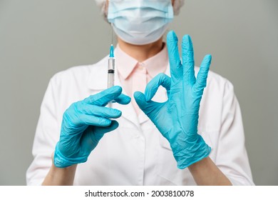 Female Doctor Or Scientist In White Medical Gown, Blue Gloves, Green Cap And Mask Holds A Syringe In Hands On White Background. She Is Ready To Give An Injection