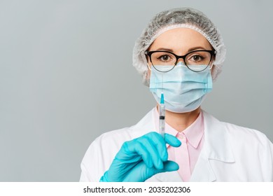 Female Doctor Or Scientist In White Medical Gown, Blue Gloves, Green Cap And Mask Holds A Syringe In Hands On White Background. She Is Ready To Give An Injection