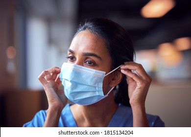 Female Doctor In Scrubs Putting On Face Mask Under Pressure In Busy Hospital During Health Pandemic