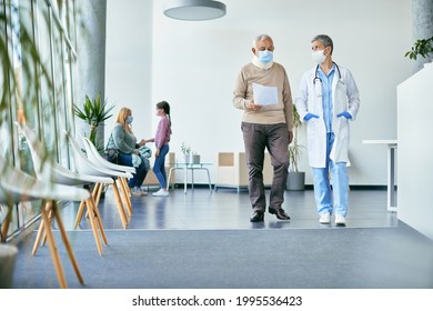 Female Doctor And Senior Man With Cervical Collar Talking About Medical Paperwork While Walking Through Lobby At Clinic And Wearing Face Masks Due To Coronavirus Pandemic.