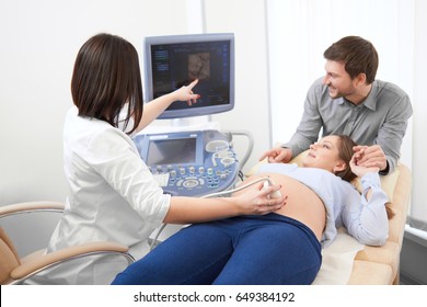 Female Doctor Showing At Screen Of Computer First Photo Of Baby In Stomach. Professional In White Using Ultrasound, Screening Pregnant Woman. Husband Carrying About Wife Of Diagnosing In Hospital.