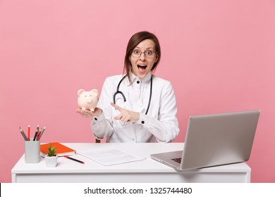 Female doctor sit at desk work on computer with medical document hold pig money in hospital isolated on pastel pink background. Woman in medical gown glasses stethoscope. Healthcare medicine concept