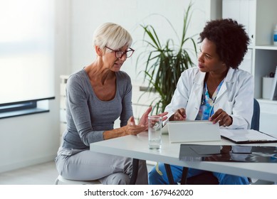 A Female Doctor Sits At Her Desk And Chats To An Elderly Female Patient While Looking At Her  Test Results