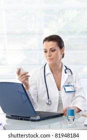 Female Doctor Sitting At Table With Laptop, Looking At Mobile Phone, Working.?