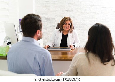 Female doctor smiling and telling a couple in their 30s about the results of medical tests. Happy gynecologist talking with a pregnant woman and her husband at her desk