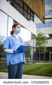 Female Doctor Standing Outside Modern Clinic Or Hospital,holding Clipboard With Patient Medical Card,wearing Blue Uniform & Protective Face Mask,nursing Home Facility Staff During Coronavirus Pandemic