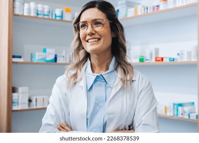 Female doctor standing in a pharmacy in a white coat. Female healthcare worker smiling with crossed arms. Woman working in a hospital dispensary. - Powered by Shutterstock