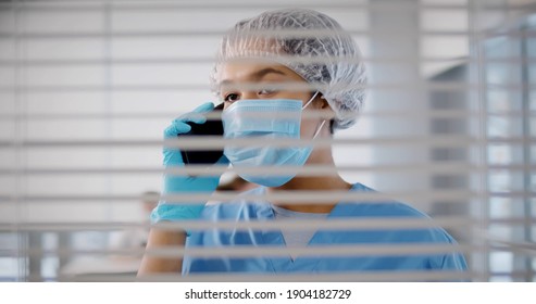 Female Doctor Talking On Mobile Phone Wearing Protective Uniform Standing Behind Window With Blinds. Afro-american Surgeon Or Nurse Having Phone Call In Hospital