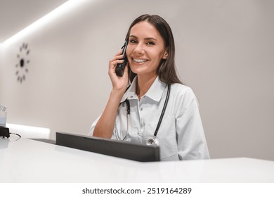 Female doctor talking with patient by phone at reception desk scheduling appointments in clinic using modern technology for communication looking at camera and smiling. Medicine health care concept - Powered by Shutterstock