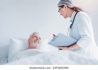 Female doctor talking to senior male patient in hospital bed. Nurse general practitioner attending elderly patient in hospital hospice. Caregiving - Powered by Shutterstock