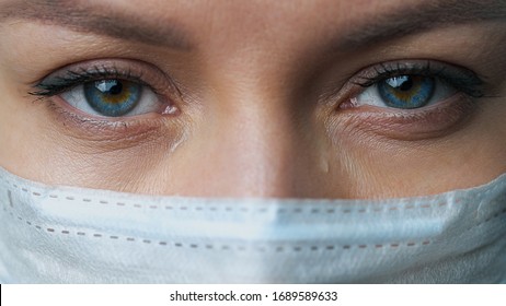 A female doctor therapist in a white robe, mask and gloves. Face close-up. The doctor cries and prays. Tears in eyes. Regret, anxiety and despair. Pandemic and virus epidemic. Coronavirus covid-19.