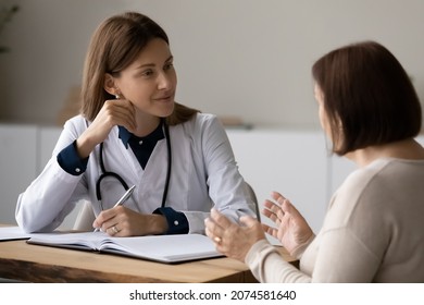 Female doctor therapist in white uniform with stethoscope listening to mature woman complaints at meeting, consulting, writing taking notes, filling patient history form, sitting at desk in hospital