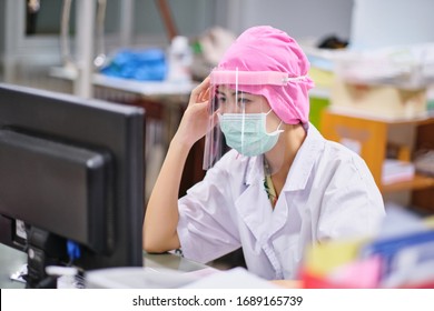 Female Doctor Under Stress And Headache Working Hard In Front Of Computer, Wearing Hospital Scrub, Face Shield Mask Protection, Concept Of Health Care Provider Front Line Against Corona Virus COVID-19