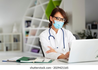 Female doctor wearing face mask while sitting at desk and using laptop at doctor’s office. 