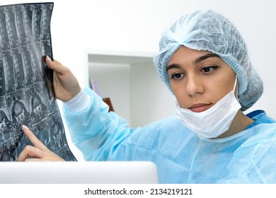 Female Doctor Wearing Face Mask And Surgical Overalls Examines An MRI Scan And Consults Online With Colleagues ​by Video Call In A Medical Office.Healthcare And Medicine Concept. 