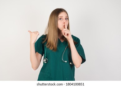Female doctor wearing a green scrubs and stethoscope is on white background asking to be quiet with finger on lips pointing with hand to the side. Silence and secret concept.