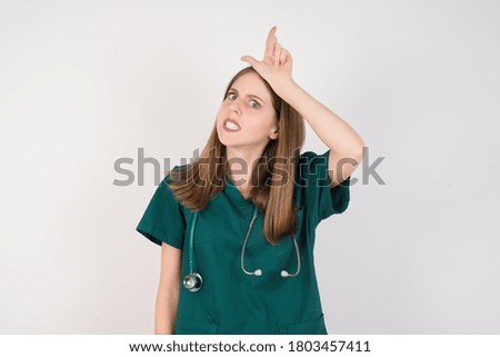 Female doctor wearing a green scrubs and stethoscope is on white background making fun of people with fingers on forehead doing loser gesture mocking and insulting.