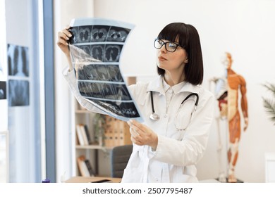 Female doctor in white coat and stethoscope examining x-ray in modern medical office. Professional healthcare worker analyzing patient scan for diagnosis and treatment planning. - Powered by Shutterstock