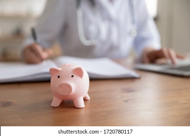 Female doctor working at desk with piggy bank box. Piggybank on table as concept of healthcare fees financial cost, money savings on health care insurance, medical care expenses concept. Close up view