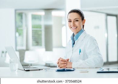 Female doctor working at office desk and smiling at camera, office interior on background