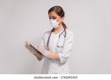 A Female Doctor Writes A Diagnosis Of The Patient. The Doctor In A Respirator And White Robe In The Clinic.