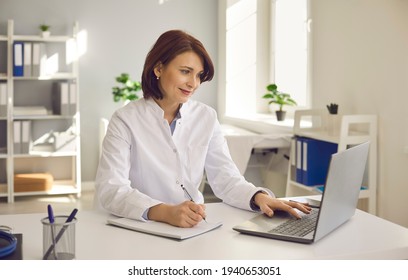 Female Doctor Writes Notes While Watching An Online Medical Webinar Or Training Seminar While Sitting With A Laptop In The Workplace. Positive Doctor Does His Best To Provide Quality Medical Care