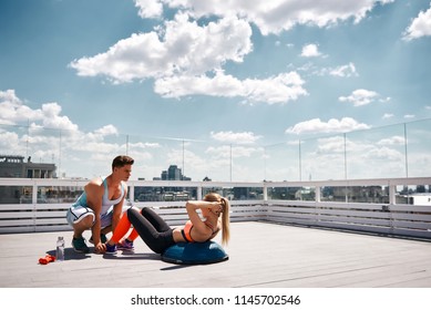 Female is doing crunches on BOSU ball while her male friend is holding her feet. They are having joint workout with sport equipment on top of urban high building. Copy space in right side - Powered by Shutterstock