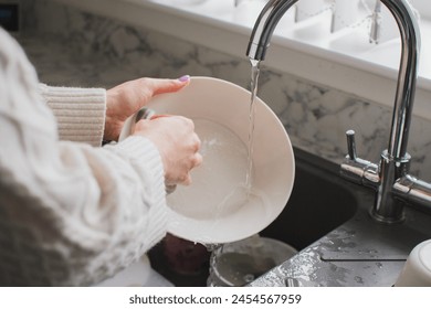 A female doing dishes, washing up in kitchen sink, closeup view. Cleaning chores. Dishwashing: washing a white plate under running water from the faucet. Cleaning the house, home routine. cleaning up - Powered by Shutterstock