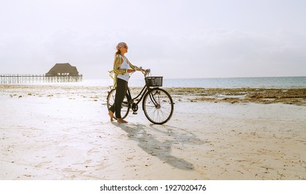 Female dressed light summer clothes have morning walk with old vintage bicycle with front basket on the lonely low tide ocean white sand coast on Kiwengwa beach on Zanzibar island, Tanzania. - Powered by Shutterstock