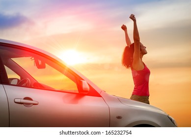 Female driver beside car raising arms and feeling the freedom of driving towards the sunset.  Woman and vehicle on beautiful sunshine background.