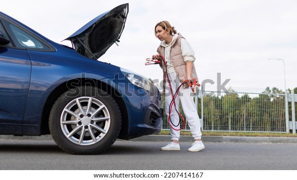 female driver at the open hood of a car with\
wires for charging the\
battery