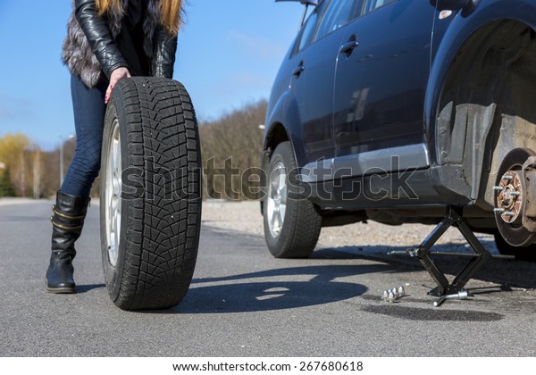 Female driver\
repairs car. Body of young female, dressed biker style, rolling big\
wheel towards broken\
car.