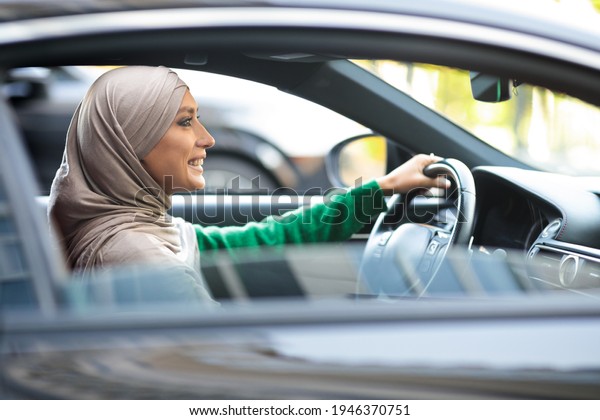 Female Driver.\
Side view profile portrait of positive islamic lady wearing\
headscarf driving her new luxury car. Smiling muslim woman in hijab\
holding steering wheel, view from\
outside