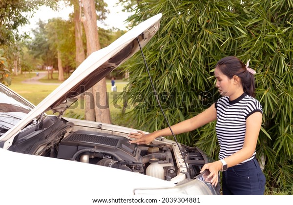 Female driver standing on street near\
her car with popped up hood looking at broken\
engine.