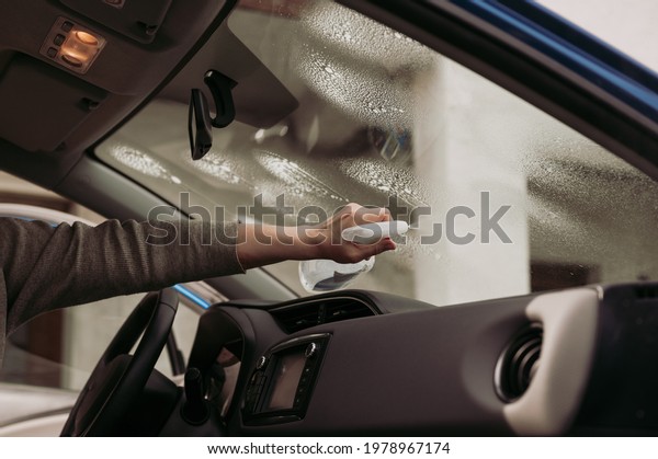 Female driver taking care of car interior hygiene.\
Woman\'s hand spraying and cleaning inside of windscreen in modern\
vehicle. 